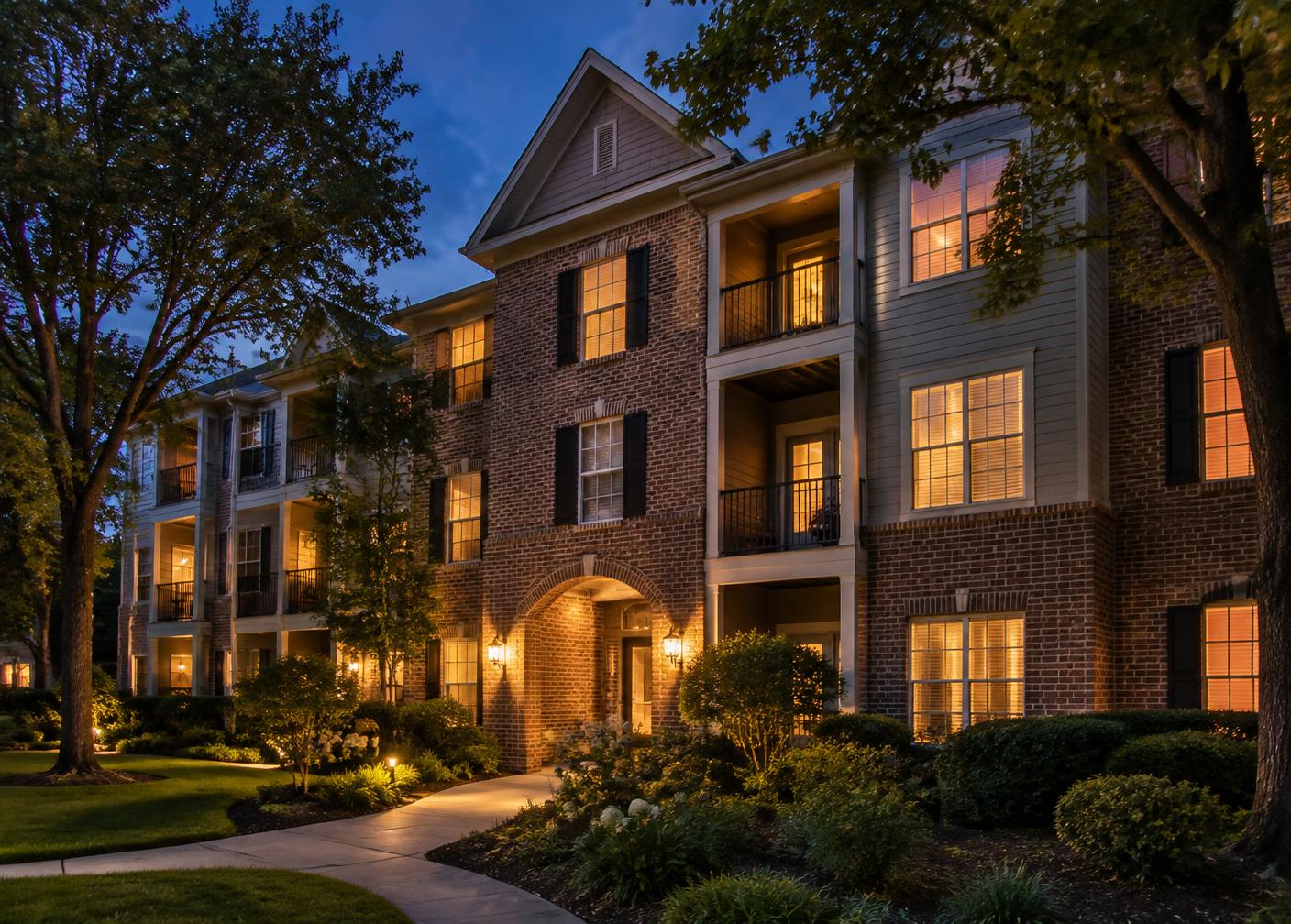 A brick multifamily community at dusk with warmly lit windows and landscaped walkways